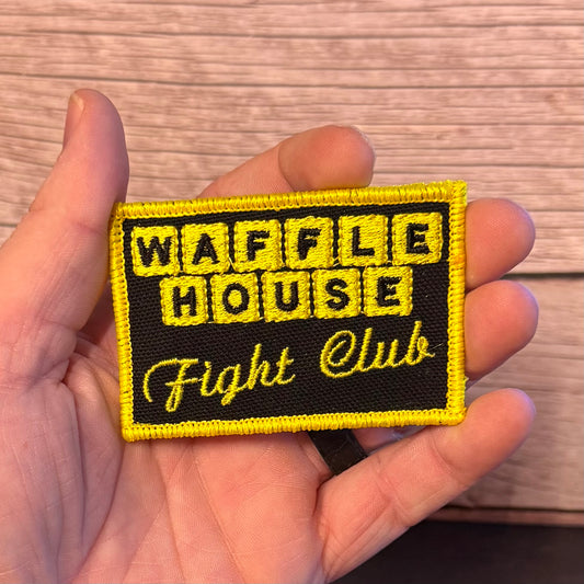A rectangular embroidered patch held in a hand, featuring a black background with yellow border and text reading 'WAFFLE HOUSE Fight Club' in yellow lettering.