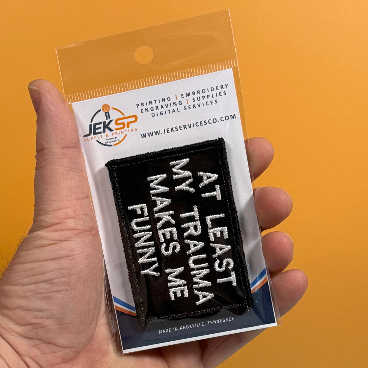A hand holds a packaged embroidered patch against a solid orange background. The patch is black with white text reading 'AT LEAST MY TRAUMA MAKES ME FUNNY.