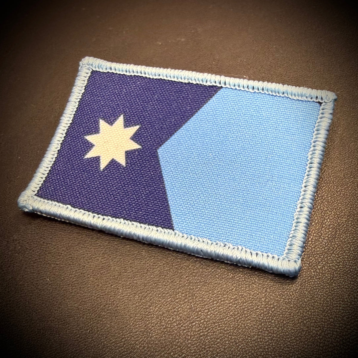 A rectangular embroidered patch featuring a diagonal split design with dark purple on the left and light blue on the right, plus a cream-colored multi-pointed star in the upper left. The patch has a light blue stitched border and is photographed on a textured brown surface.