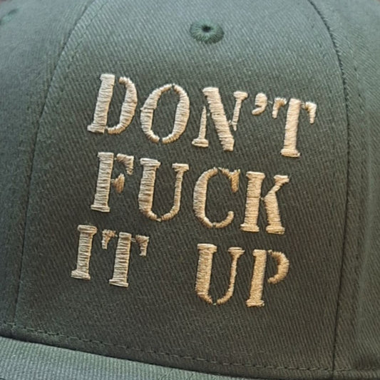 Green cap with gold text 'DON'T FUCK IT UP' on a wooden surface