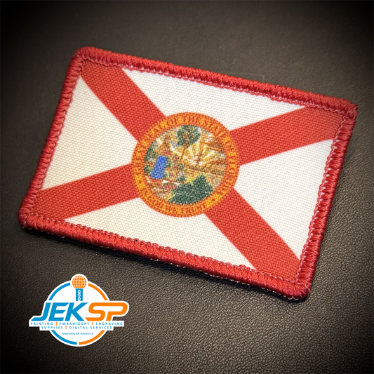 An embroidered rectangular patch of the Florida state flag, featuring a red saltire on a white background with the state seal in the center, photographed on a dark textured surface. A 'JEKSP' watermark is visible in the bottom-left corner.