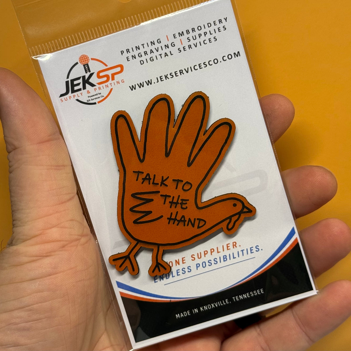 A hand holding a packaged orange turkey-shaped enamel pin with the text 'TALK TO THE HAND' on a white backing card from JEK Supply & Printing.