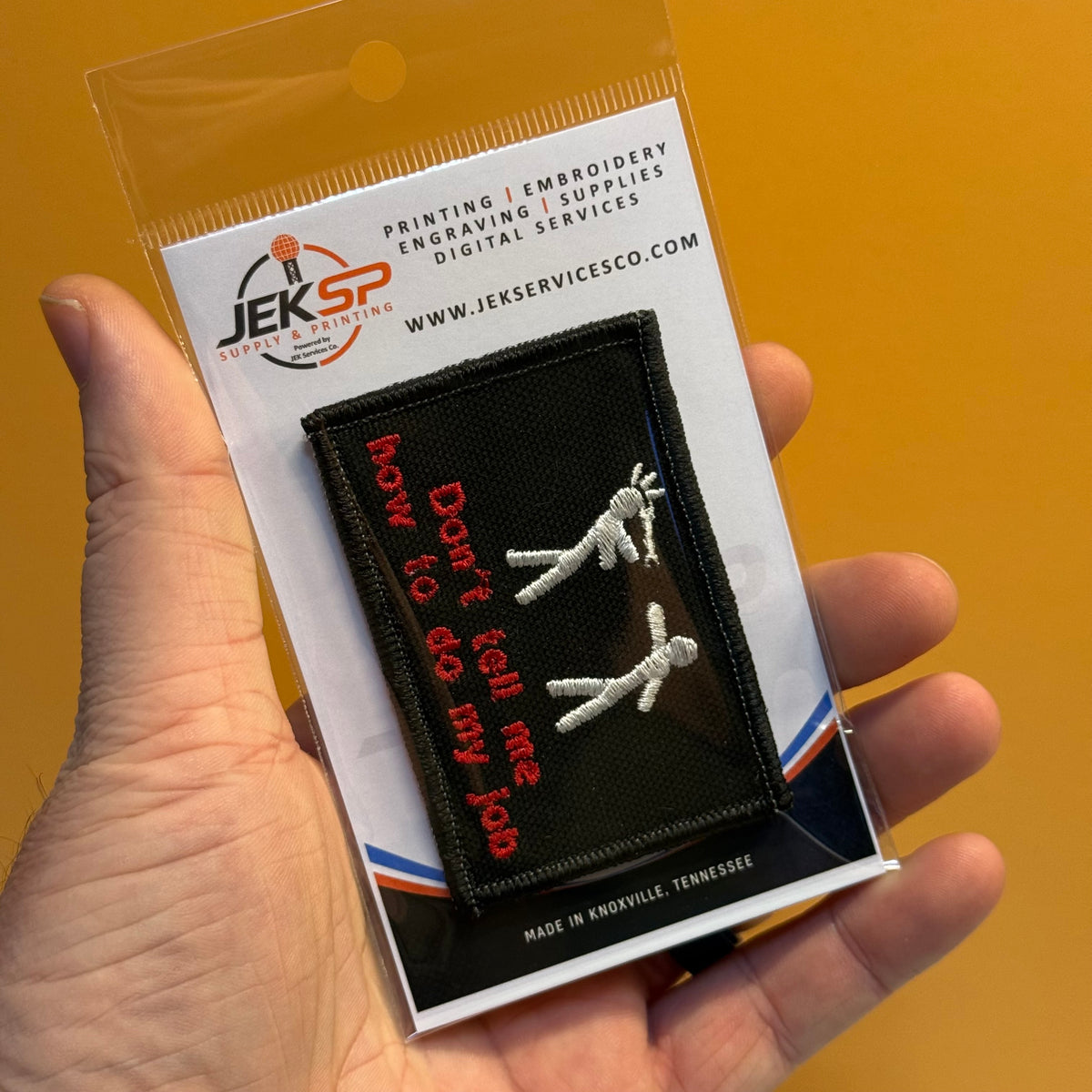 A hand holds a clear plastic blister pack containing a black embroidered patch with red text reading 'Don't tell my job' and white stick-figure-like embroidery. The backing card shows the JEKSP logo and website text against a warm tan background.