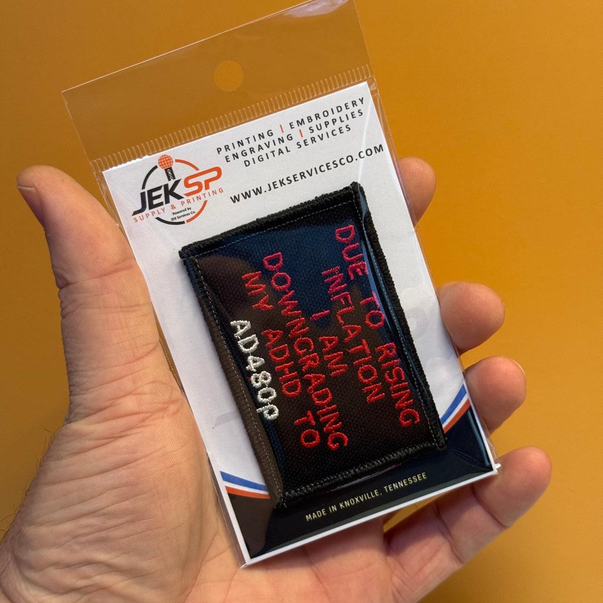 A hand holds a clear plastic blister pack containing a rectangular embroidered patch. The patch has a black background with red and white text reading, 'DUE TO RISING INFLATION I AM A ADDICT TO DOWNING MY ADDICTION TO AD4809.