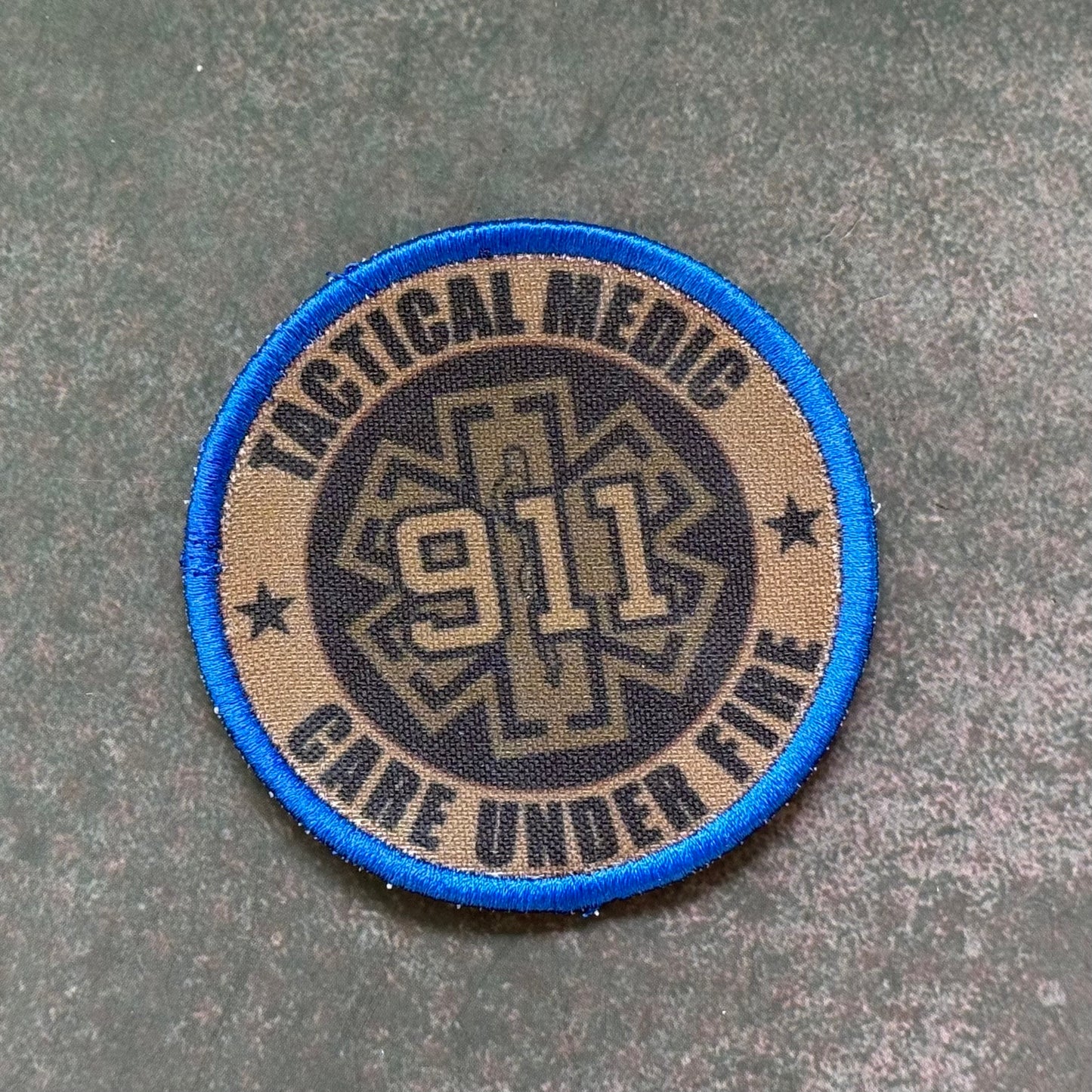 A circular tactical medic patch with a blue border, tan background, and a dark center featuring the text 'TACTICAL MEDIC', '911', and 'CARE UNDER FIRE' on a textured gray surface.