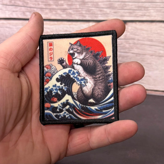 A hand holding a square embroidered patch featuring a cat in a Godzilla-style pose riding a blue wave with a red sun and Japanese text, set against a light wood background.