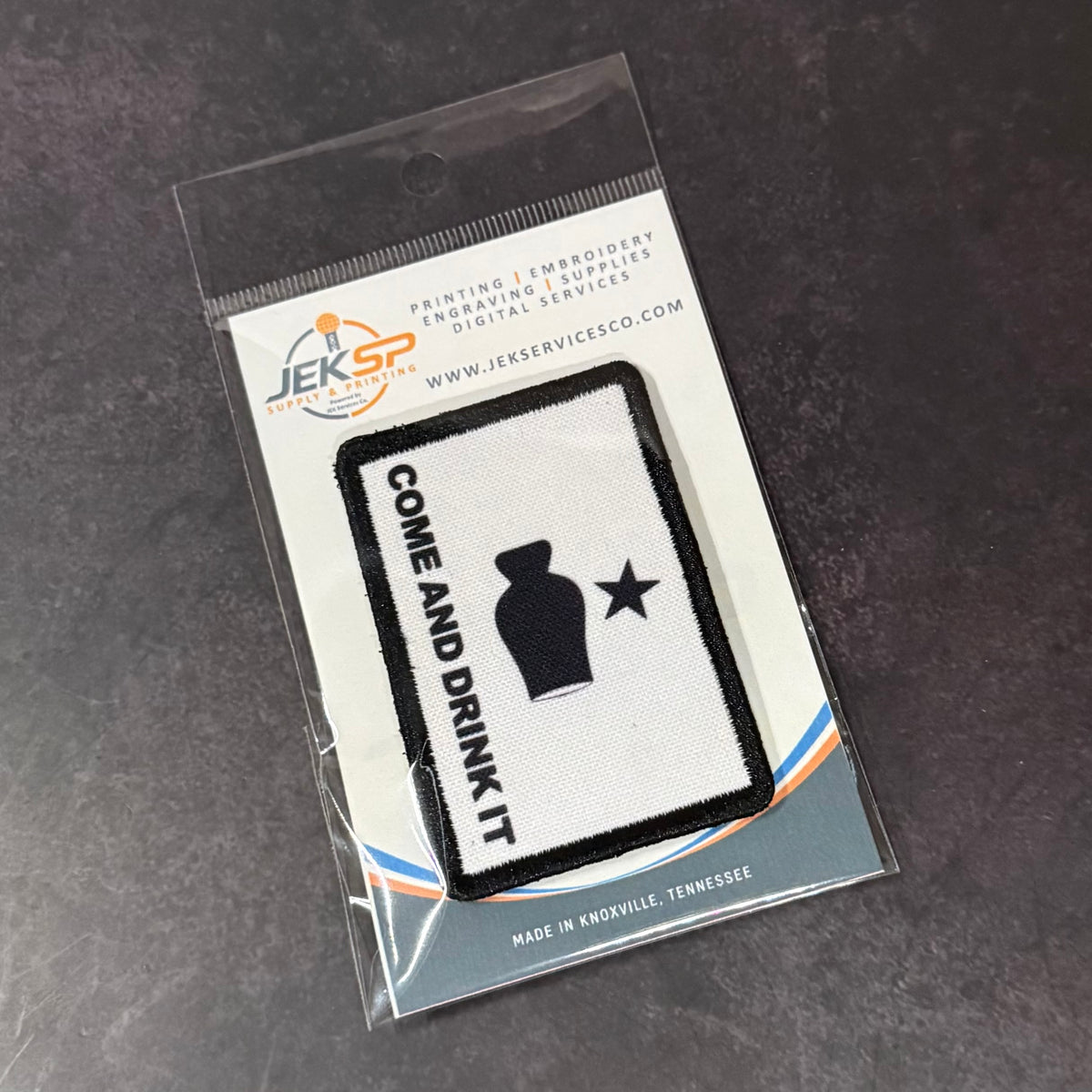 Bourbon Whiskey Come and Drink It printed canvas morale patch, in clear plastic packaging on a dark textured surface. The patch is white with a black border, featuring the design lettering alongside a black silhouette of a.