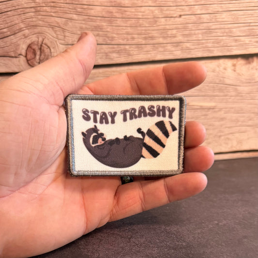 A hand holding a rectangular embroidered patch that reads “STAY TRASHY” above a sleeping raccoon with a striped tail, set against a rustic wood plank background.