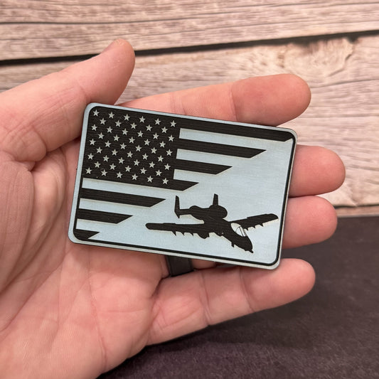 A hand holding a rectangular metal badge featuring a black and white American flag design with a silhouette of a fighter jet, set against a rustic wood background.