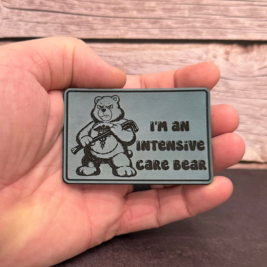 A hand holding a rectangular gray patch featuring a cartoon bear holding a thermometer and the text 'I'M AN INTENSIVE CARE BEAR', set against a rustic wood plank background.