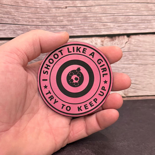 A hand holding a round pink and black sticker with the text 'I SHOOT LIKE A GIRL TRY TO KEEP UP' and a target graphic in the center, set against a rustic wood plank background.