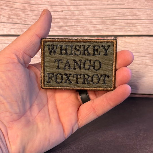 A rectangular embroidered tactical patch with the words 'WHISKEY TANGO FOXTROT' in black lettering on an olive drab background, held in a person's hand against a light wood plank background.