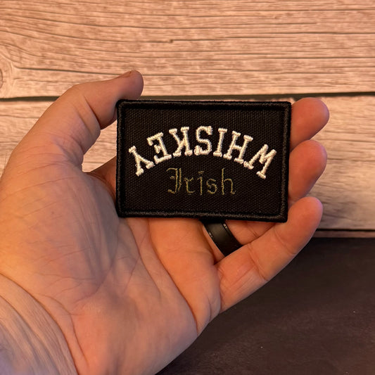 A hand holding a rectangular black embroidered patch with the words 'Whiskey Irish' in white and gold lettering, set against a rustic wooden background.
