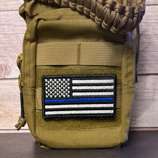 An olive drab tactical pouch with a 'Thin Blue Line' American flag patch on the front, featuring a paracord strap at the top, set against a rustic wood background.