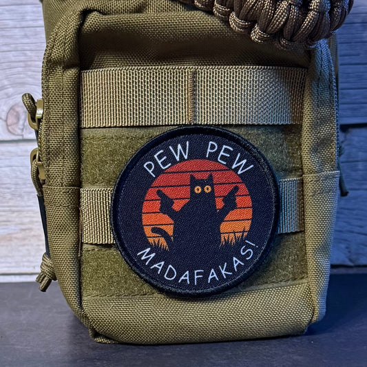 An olive green tactical pouch with a circular embroidered patch featuring a black cat silhouette against an orange sunset and the text 'PEW PEW MADAFAKASI'.