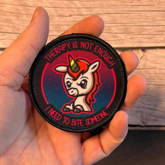 A hand holding a round embroidered patch featuring a cartoon unicorn with a red horn and the text 'THERAPY IS NOT ENOUGH I NEED TO BITE SOMEONE' against a wood plank background.