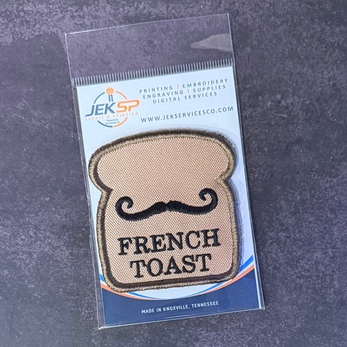French Toast embroidered morale patch: shaped like a slice of French toast with a black mustache and the words 'FRENCH TOAST' in black, sealed in a clear plastic package with a blue and white Jeksp Service Co. backing.