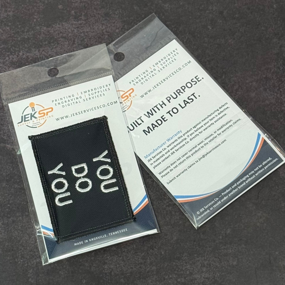A black rectangular embroidered patch with the words 'you do you' in white, sealed in a clear plastic package with a white backing card. A second white card with printed text is placed behind it on a dark textured surface.