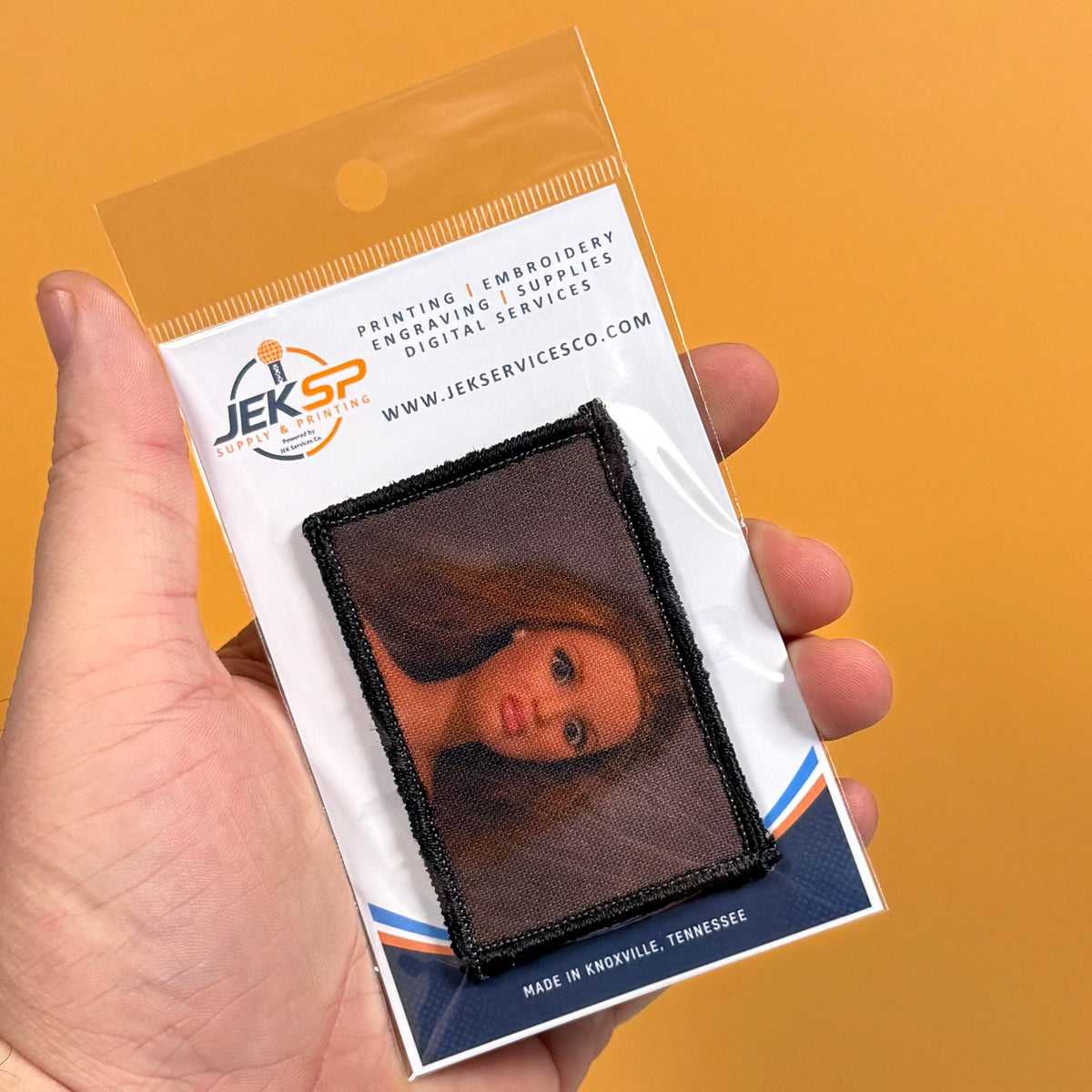 A hand holds a clear plastic package containing a square embroidered patch with a doll's face on a dark mesh background. The package card shows the brand name JEKSP Industries, Inc.