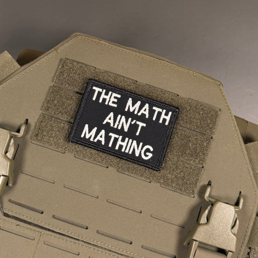 The Math Ain't Mathing embroidered morale patch: black tactical patch with white embroidered text reading 'THE MATH AIN'T MATHING' attached to tan MOLLE webbing on a tan tactical vest or plate carrier.