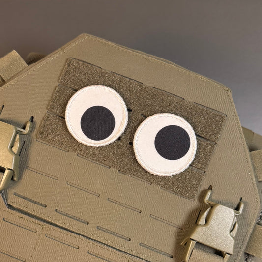 A close-up of a tan tactical vest or plate carrier with two circular googly-style eyes attached on a hook-and-loop panel.