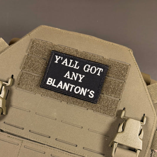 Got Any Blanton's embroidered morale patch: tactical plate carrier vest in coyote tan, featuring a black rectangular embroidered patch with the white text 'Y'ALL GOT ANY BLANTON'S' mounted on hook-and-loop fabric.