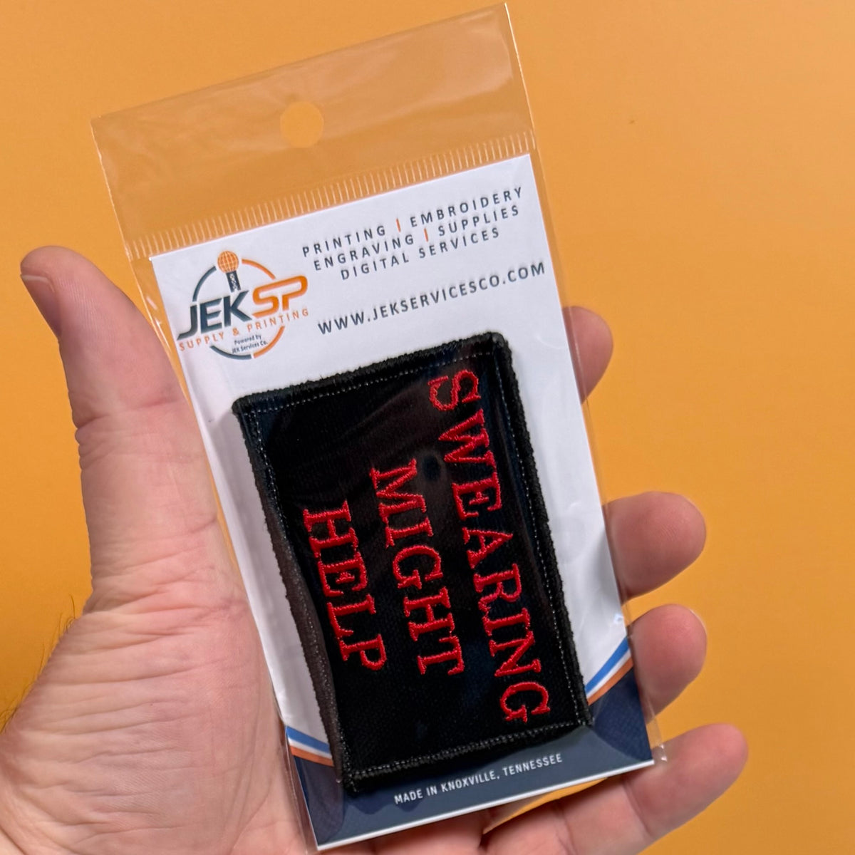A hand holding a clear plastic blister pack containing a black rectangular patch with red embroidered text reading 'SWEARING MIGHT HELP'. The backing card shows the JKS Service Co.