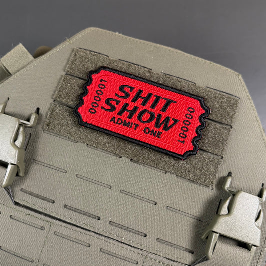 Welcome To The Shit Show Ticket embroidered morale patch: red embroidered patch with black text reading 'SHIT SHOW ADMIT ONE' and '100000' on both sides, mounted on a tan tactical vest with a hook-and-loop patch panel.