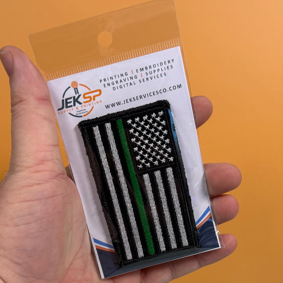 Thin Green Line Tactical American Flag embroidered morale patch: hand holds a packaged embroidered patch featuring a black-and-white American flag with a single green stripe, on a white backing card from JEKSP Supplies & Engraving..