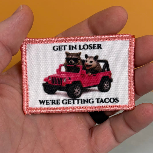 A hand holding a rectangular embroidered patch with a pink border. The patch features a white background with the text 'GET IN LOSER WE'RE GETTING TACOS' in black, surrounding an illustration of two raccoons sitting in a red jeep.