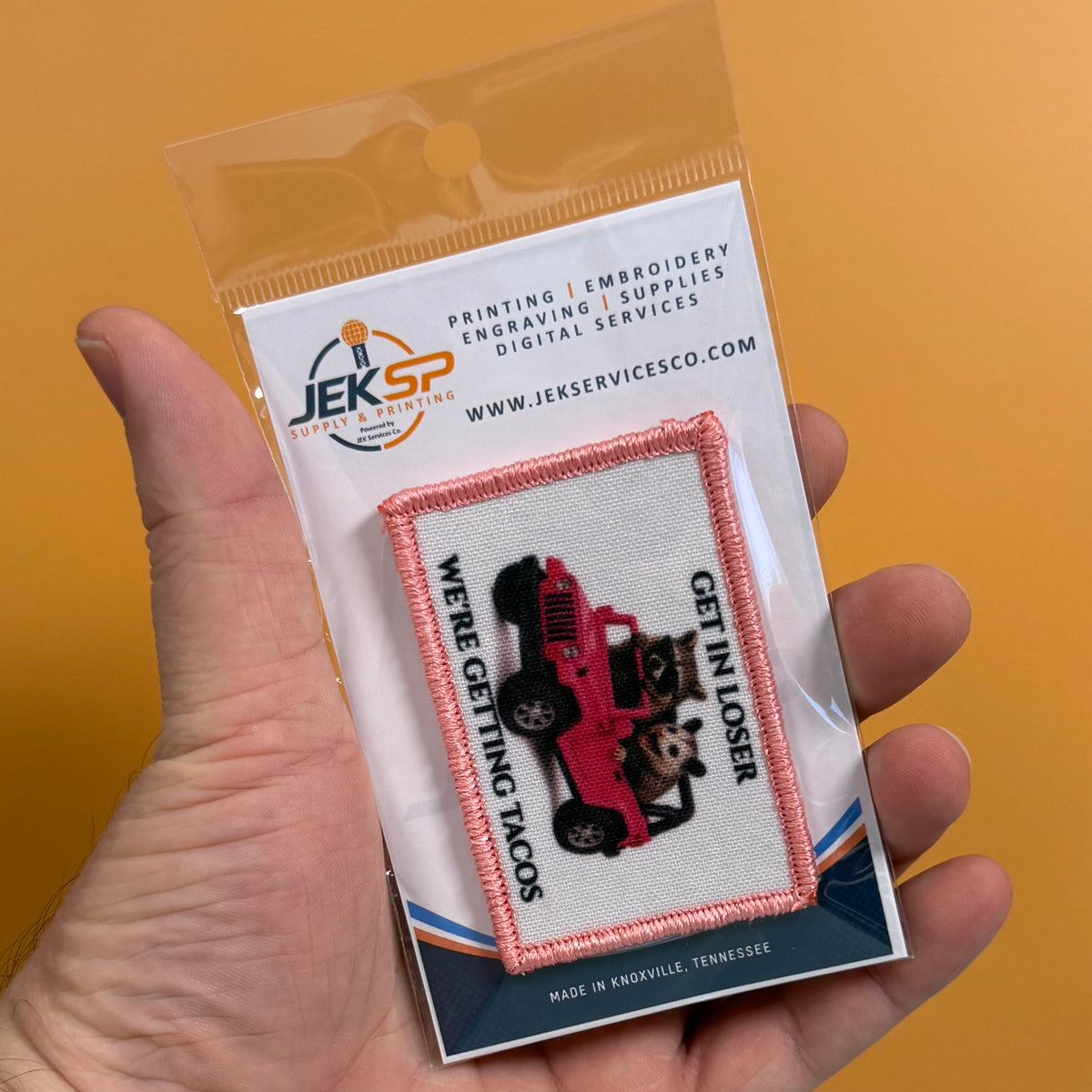 Get In Loser We're Getting Tacos printed canvas morale patch, a hand holds a clear plastic blister pack containing an printed patch. The patch shows a red jeep with a dog and cat inside, surrounded by pink stitching and the.