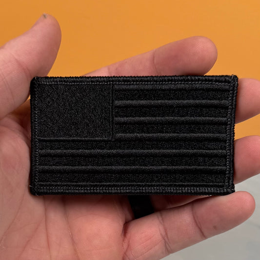 A black embroidered American flag patch held in a hand against a solid orange background.