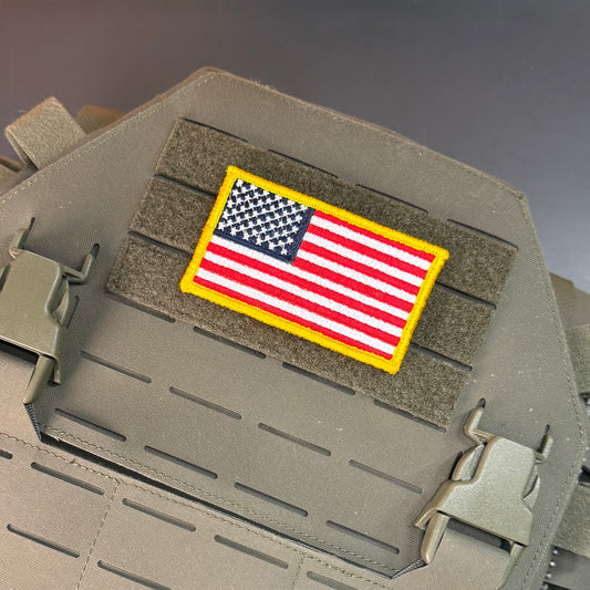 A rectangular embroidered American flag patch with a yellow border, mounted on a tan tactical vest or plate carrier with hook-and-loop fastening.