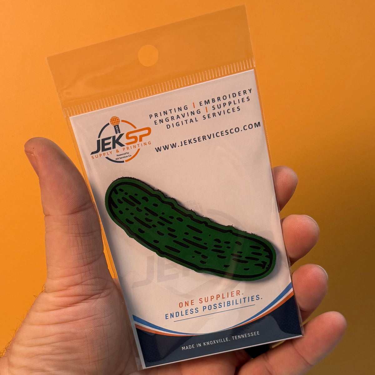 Dill Pickle laser-engraved synthetic morale patch: a hand holding a packaged dark green pickle-shaped patch on a white backing card, with an orange background. The card shows the Jeksp Service Co.