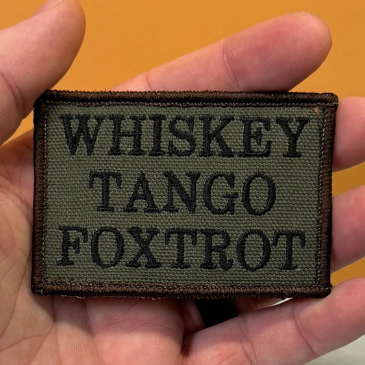 A rectangular embroidered patch held in a hand, featuring the text 'WHISKEY TANGO FOXTROT' in black on an olive drab background with a dark brown border.