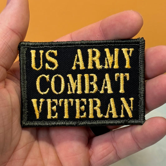 A rectangular embroidered patch reading 'US ARMY COMBAT VETERAN' in gold lettering on a black background, held in a person's hand against an orange background.