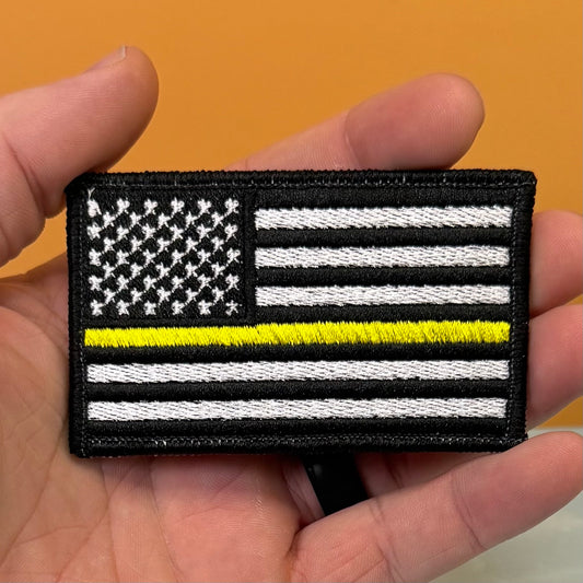 A hand holding a rectangular embroidered patch featuring a black-and-white American flag design with a single bright yellow horizontal stripe, shown against an orange background.
