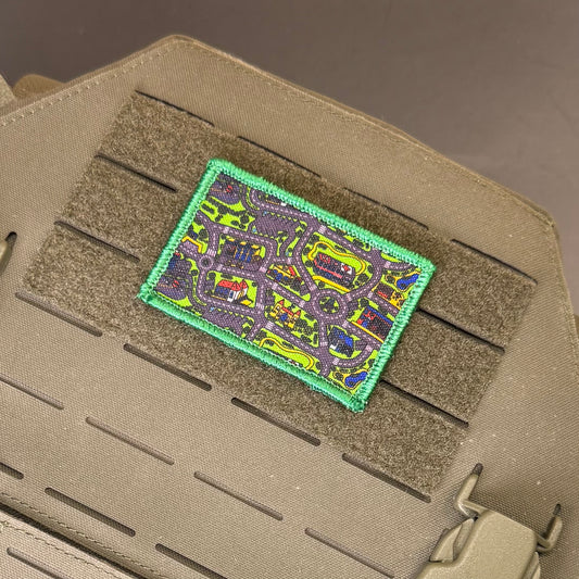 A rectangular tactical patch featuring a colorful street map design with a bright green border, shown attached to a tan tactical vest with Velcro loops.