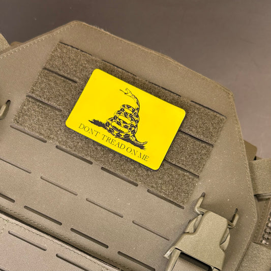 A bright yellow rectangular tactical patch featuring a black coiled rattlesnake and the text 'DON'T TREAD ON ME' attached to olive drab MOLLE webbing on a tactical vest.