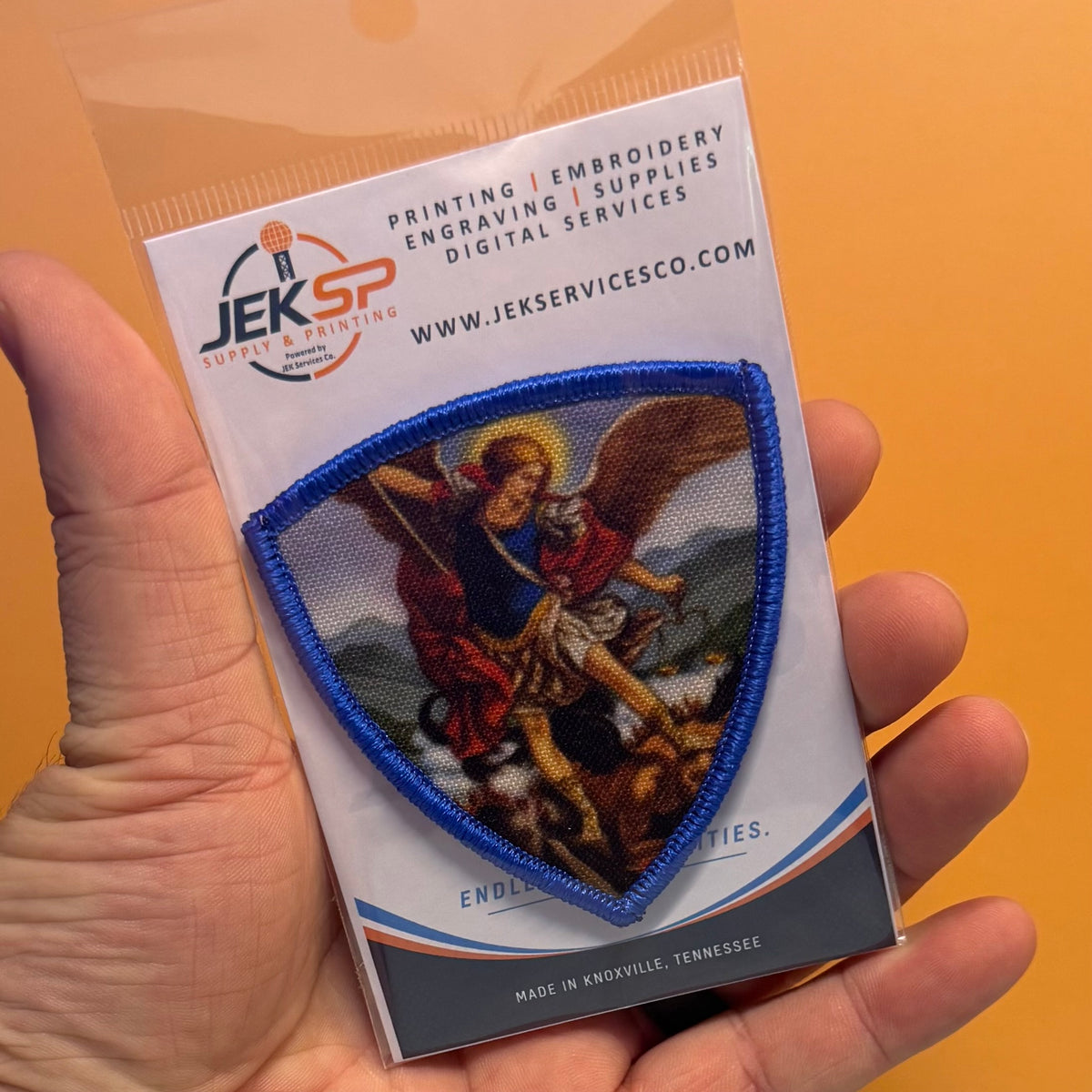 Saint Michael Shield printed canvas morale patch, a hand holds a packaged printed patch featuring a blue-bordered shield with a religious scene. The backing card shows the Jekspaint logo and text for printing, embroidery.