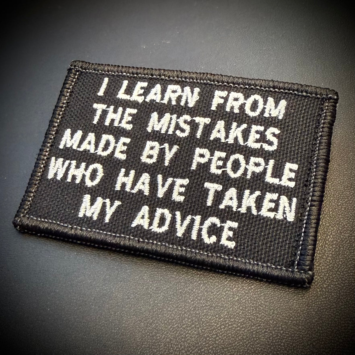 A rectangular black embroidered patch with white text reading 'I LEARN FROM THE MISTAKES MADE BY PEOPLE WHO HAVE TAKEN MY ADVICE', shown on a dark textured background.