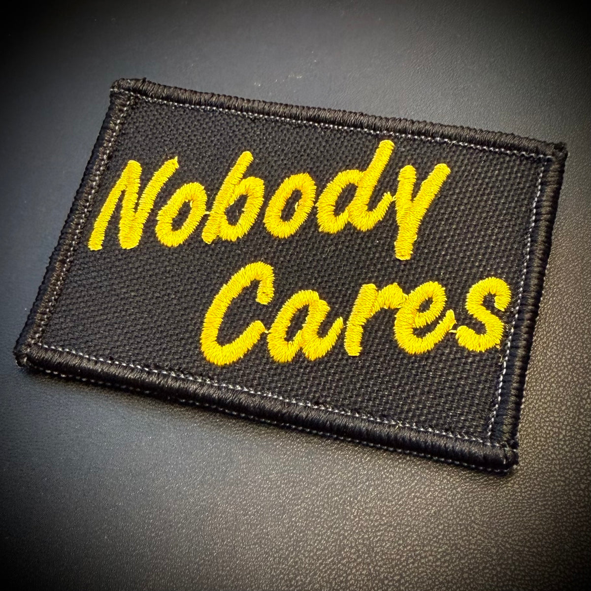 A rectangular black embroidered patch with the phrase 'Nobody Cares' in bright yellow script, shown on a dark textured surface.