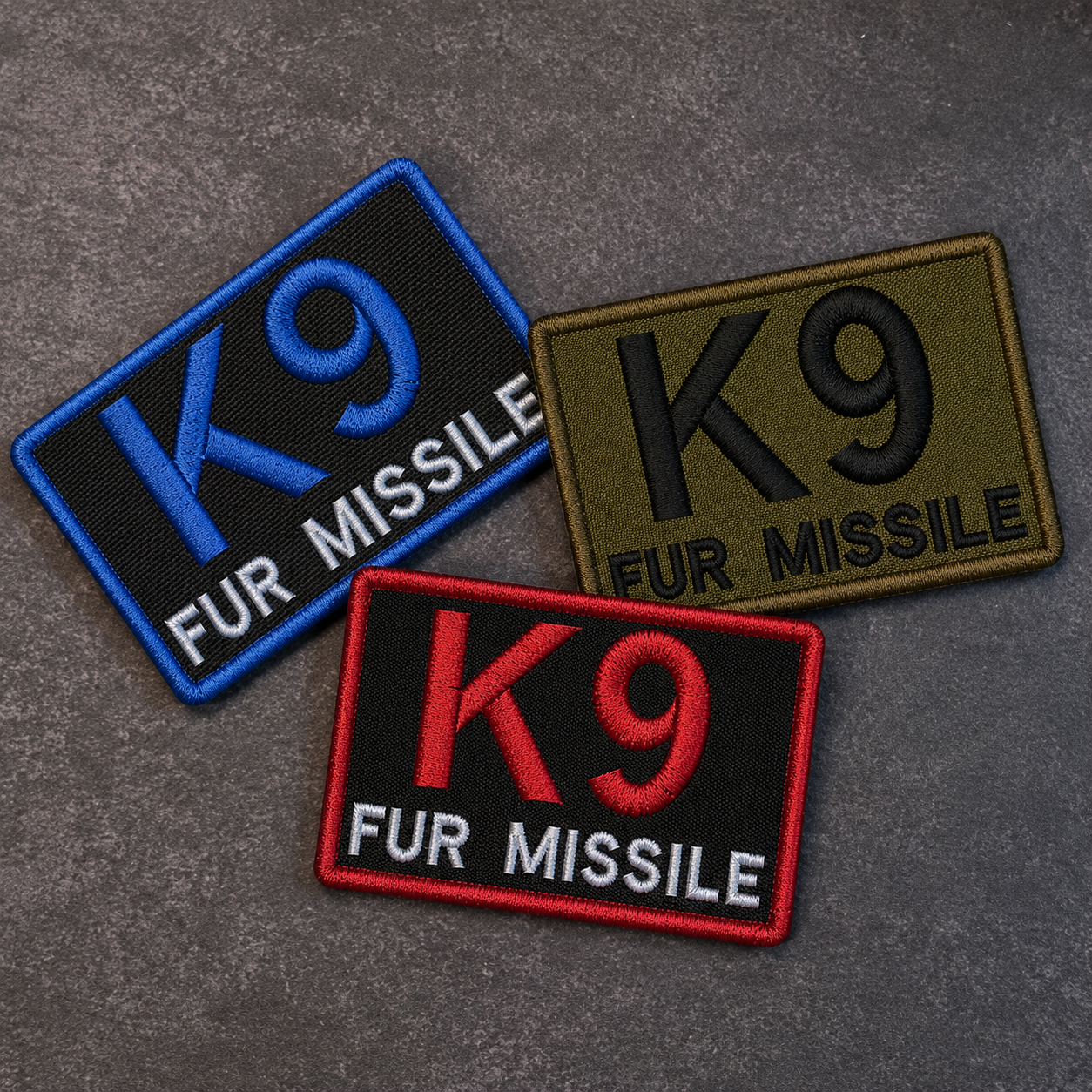 Three embroidered rectangular patches with 'K9' and 'FUR MISSILE' text, shown on a gray textured background. One patch is blue and black, one is olive green and black, and one is red and black.