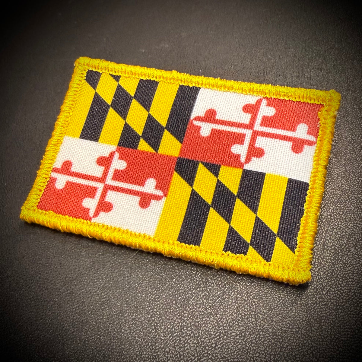 A rectangular embroidered patch of the Maryland state flag, featuring its distinctive black and yellow checkered quadrants with red and white crosses, displayed on a dark textured surface.