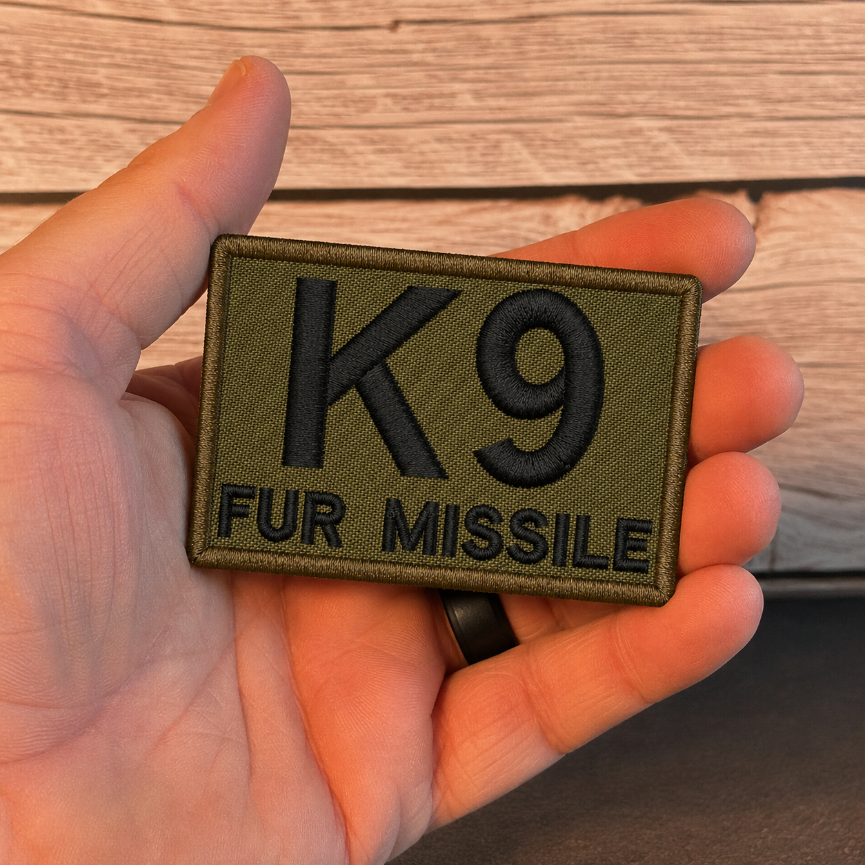 An olive drab green embroidered tactical patch with black text reading 'K9 FUR MISSILE', held in a person's hand against a rustic wooden background.