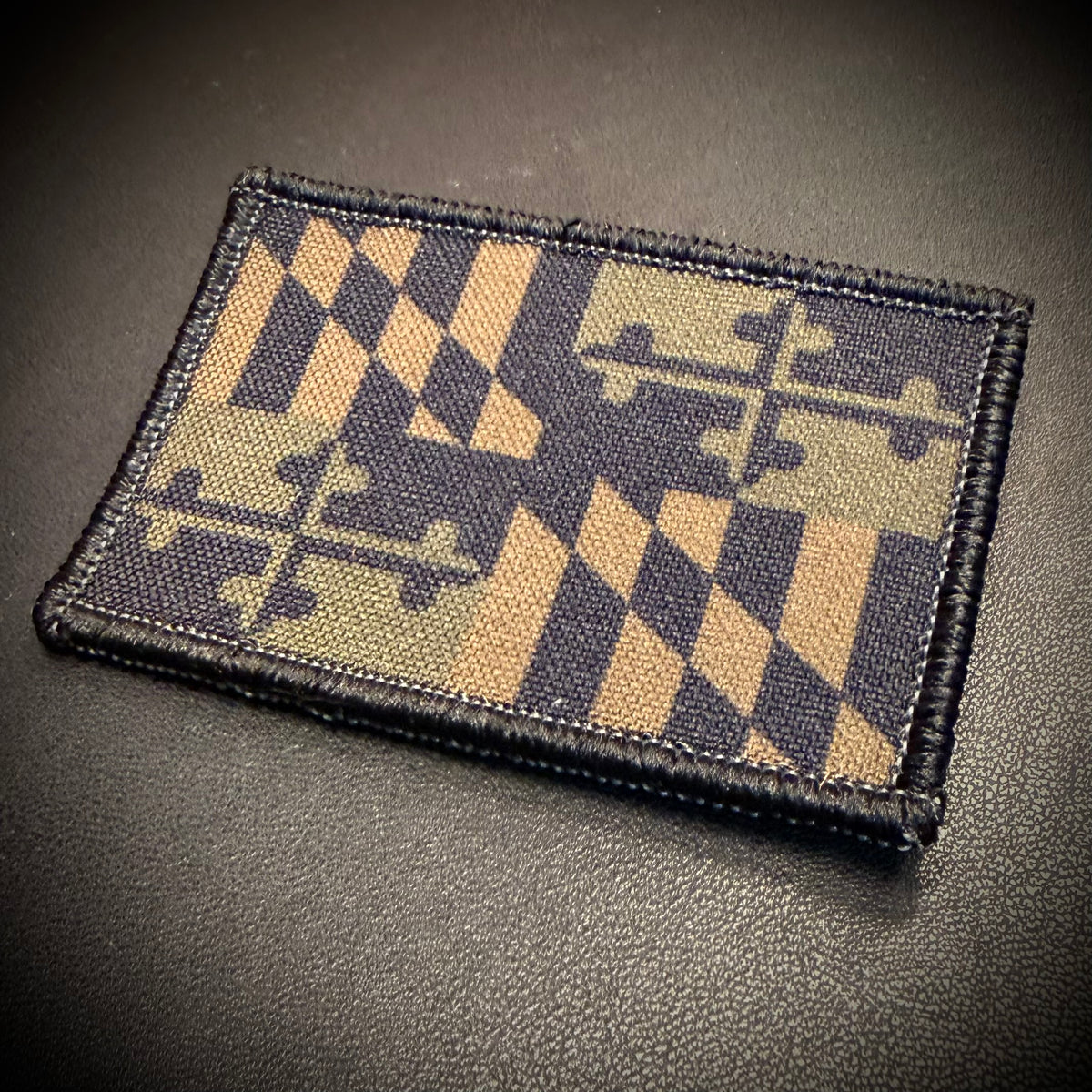 A rectangular tactical patch featuring a black and tan puzzle-piece diamond pattern, bordered in black and placed on a dark textured surface.