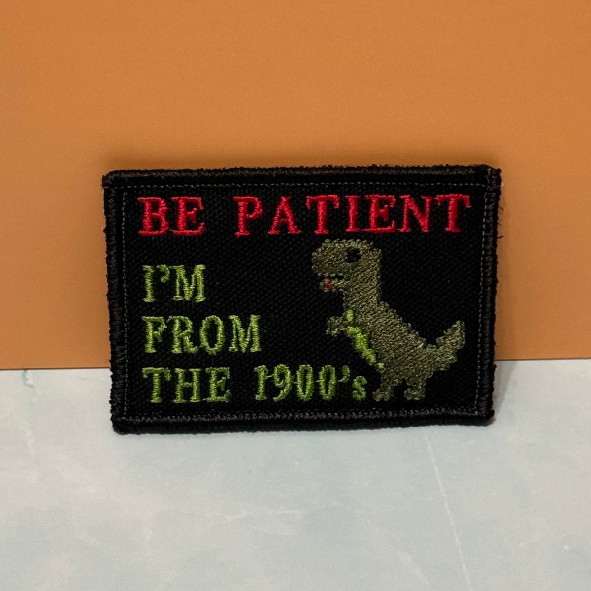 A rectangular embroidered patch with a black background featuring the text 'BE PATIENT I'M FROM THE 1900's' in red and green lettering, alongside a green dinosaur illustration, shown against an orange and white background.