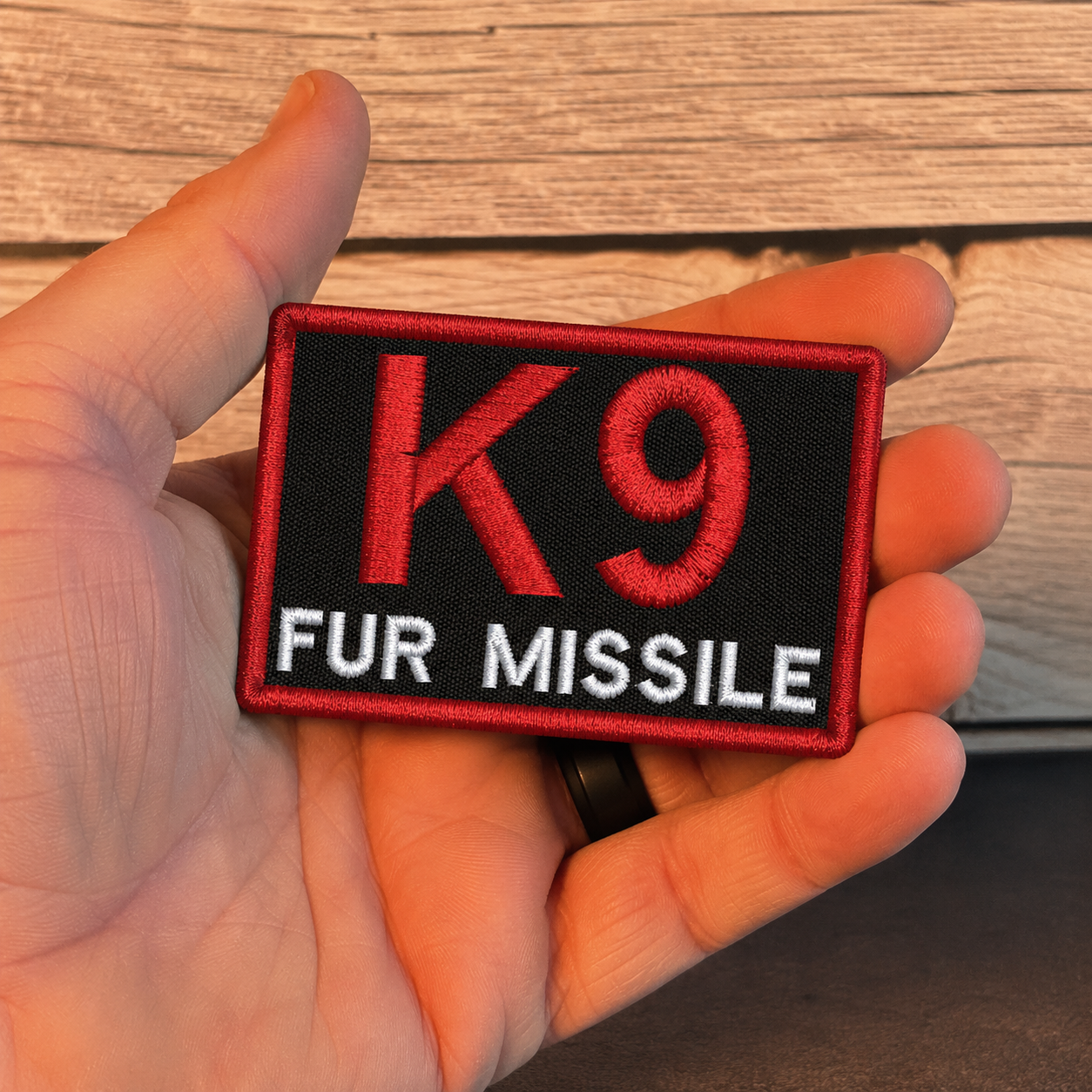 A hand holding a rectangular embroidered patch with a black background, red border, large red 'K9' text, and white 'FUR MISSILE' text, against a wood plank background.