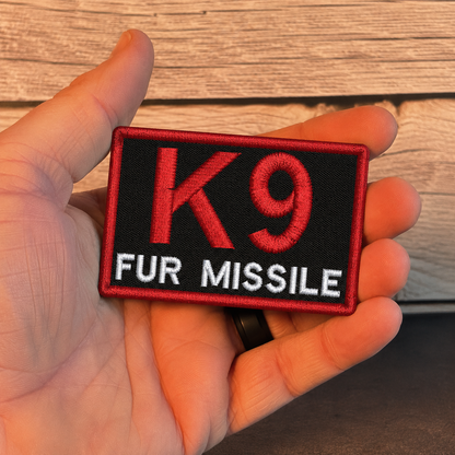 A hand holding a rectangular embroidered patch with a black background, red border, large red 'K9' text, and white 'FUR MISSILE' text, against a wood plank background.