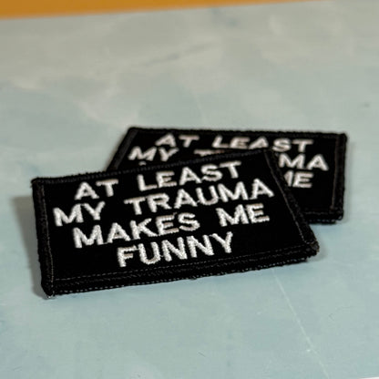 Two rectangular embroidered patches with black backgrounds and white text reading 'AT LEAST MY TRAUMA MAKES ME FUNNY,' photographed on a pale blue surface.